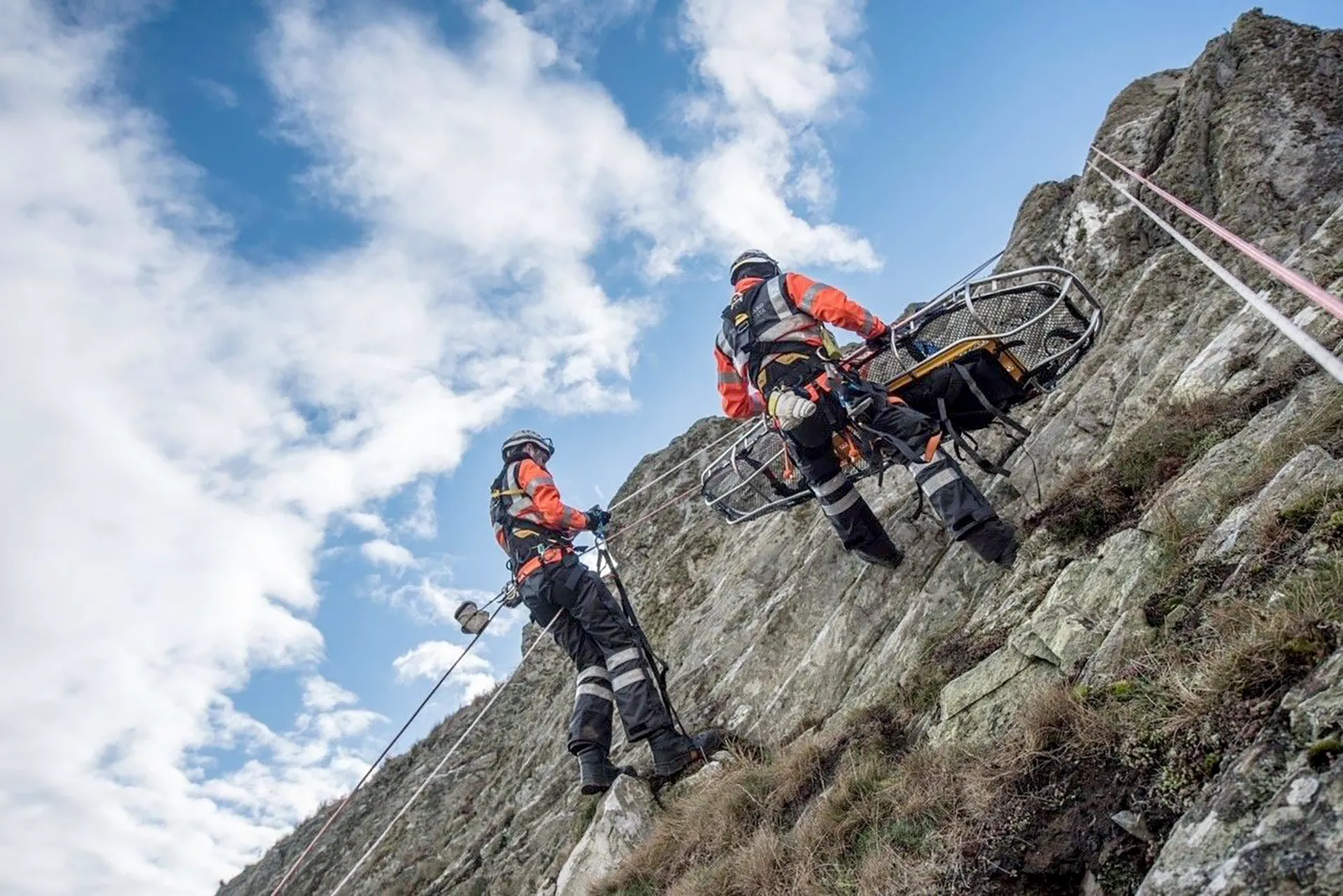 Two rescue personnel using ropes to manoeuvre a stretcher along a steep rocky cliff during a technical rescue operation