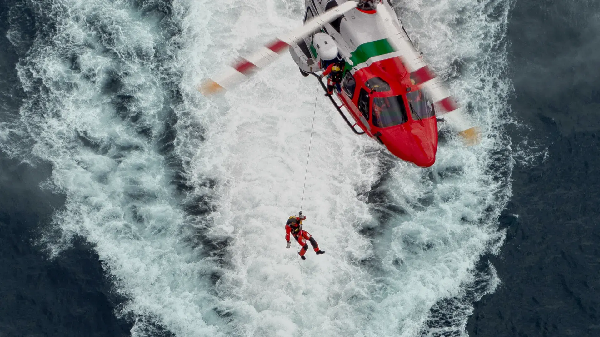 A rescue helicopter hovering above rough sea conditions while a crew member is being lowered on a winch line