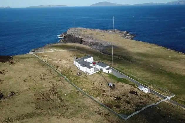 Aerial view of a remote coastal facility with buildings, access road, and communications masts overlooking the sea